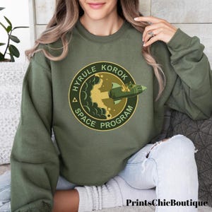 Peut inclure: Un sweat-shirt vert avec un graphique circulaire représentant une fusée décollant avec le texte "Hyrule Korok Space Program".