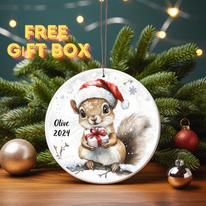 May include: A white ceramic Christmas ornament featuring a cartoon squirrel wearing a red Santa hat and holding a gift box with a red ribbon. The ornament has the name "Olive" and the year "2024" written on it.