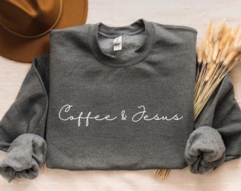 May include: A dark gray sweatshirt with the words "Coffee & Jesus" in a white script. A brown hat and dried wheat stalks are in the background. The text "PRINTSCHICBOUTIQUE" is at the bottom.