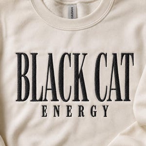 Puede incluir: Una sudadera de cuello redondo color crema con texto bordado en negro en el pecho que dice BLACK CAT ENERGY. La prenda tiene un cuello acanalado y una etiqueta de la marca Gildan visible en el cuello interior, con un diseño sencillo.