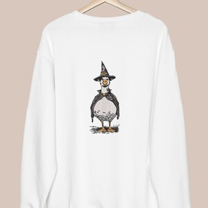 May include: White sweatshirt featuring a whimsical illustration of a goose dressed as a witch. The goose wears a black witch hat with a gold buckle and a black cape. The design is centered on the front of the sweatshirt, perfect for Halloween.