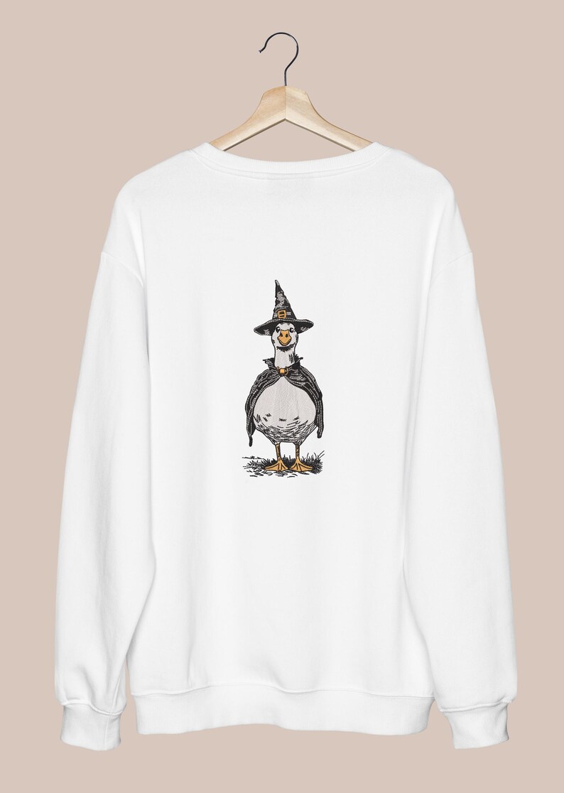 May include: White sweatshirt featuring a whimsical illustration of a goose dressed as a witch. The goose wears a black witch hat with a gold buckle and a black cape. The design is centered on the front of the sweatshirt, perfect for Halloween.