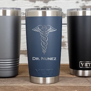 May include: Three stainless steel insulated tumblers with silver lids. One is gray, one is navy blue with a medical symbol and "Dr. Nunez" text, and one is black with the "YETI" logo. All have a silver base.