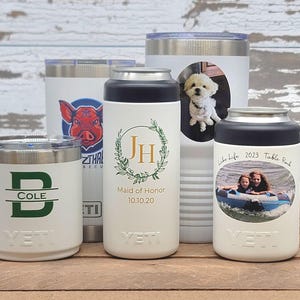 May include: A collection of white Yeti tumblers and can coolers with various custom designs. One features a pig graphic, another a dog photo, and others include initials, names, and dates. The tumblers are on a wooden surface.