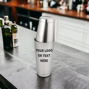 May include: A stainless steel cocktail shaker with a lid, resting on a bar. The shaker has the text "YOUR LOGO OR TEXT HERE" printed on the front. Two bottles are visible in the background.