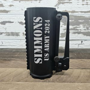 May include: Black mug with a unique handle design, featuring the text "SIMMONS US ARMY 2024" in white. The mug has a textured surface and is set against a wooden background.