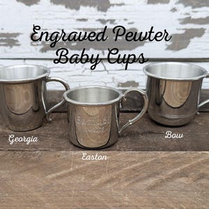 May include: Three engraved pewter baby cups with handles. The cups are silver and have different designs, with the names "Georgia", "Easton", and "Bow" written below them. The background is a weathered white wooden surface.