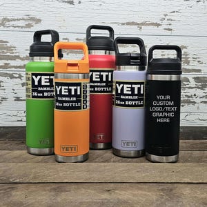 May include: Five insulated water bottles in various colors: green, orange, red, lavender, and black. Each bottle has a black handle and the brand name "YETI" printed on it. The black bottle has space for a custom logo or text.