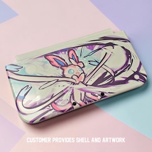 May include: A white handheld gaming console case featuring a colorful illustration of a Sylveon character. The artwork includes pink, purple, and blue accents. The text "CUSTOMER PROVIDES SHELL AND ARTWORK" is printed at the bottom.