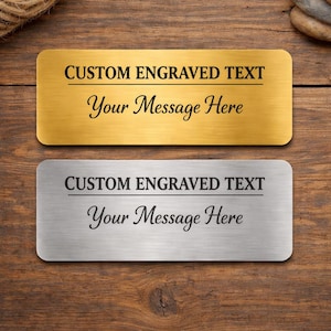 May include: Two rectangular nameplates, one gold and one silver, with rounded corners. Each plate has the text "CUSTOM ENGRAVED TEXT" above "Your Message Here" in a script font. The background is a wooden surface.