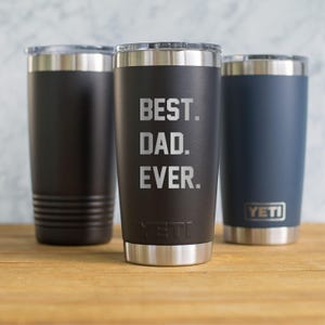 May include: Three insulated tumblers with silver rims and lids. The center tumbler is black with the text "BEST. DAD. EVER." in white. The other two tumblers are black and navy blue. The brand name "YETI" is visible on two tumblers.