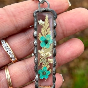 May include: A rectangular pendant necklace with dried flowers set in clear resin. The pendant has a silver-coloured metal frame with a textured edge. The flowers include small white blooms and vibrant turquoise flowers.