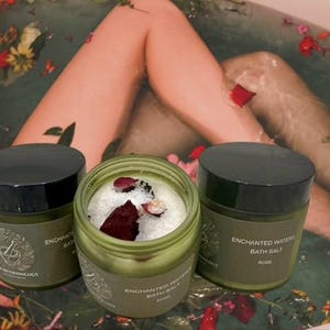May include: Three green jars of bath products with black lids. One jar is open, revealing white bath salts and rose petals. The other two jars are closed. The text "Enchanted Waters Bath Salt" is visible on the labels.