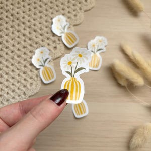Peut inclure: Un autocollant découpé représentant un vase rayé jaune et blanc contenant deux marguerites blanches à cœur jaune et tiges vertes. L'autocollant a une bordure blanche et est présenté sur une surface en bois clair avec des herbes séchées.