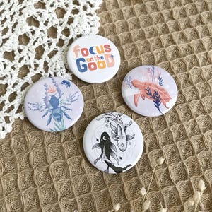 Peut inclure: Quatre badges ronds avec des motifs uniques. L'un des badges affiche le texte "Focus on the Good" en lettres colorées. Les autres présentent des illustrations d'une abeille, de poissons koï et d'une tortue de mer. Les badges sont sur une surface beige texturée.