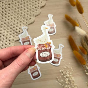 Peut inclure: Une main tenant un autocollant représentant une oie blanche perchée sur un pot de confiture étiqueté "Fraise des bois". Plusieurs autocollants similaires sont éparpillés, avec des fleurs séchées en arrière-plan. Le design de l'autocollant a une esthétique rustique et champêtre.
