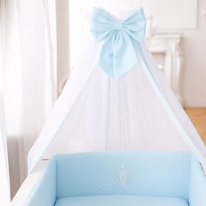 May include: A baby sleeps peacefully in a white crib with a light blue quilted bumper and mattress. A light blue bow adorns the canopy. The crib has a white frame and is in a well-lit room.