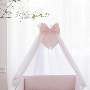 Può includere: Una culla bianca con baldacchino e biancheria da letto rosa. Il baldacchino è adornato da un grande fiocco rosa. La biancheria da letto della culla comprende un paracolpi trapuntato e un lenzuolo con angoli, entrambi rosa. La culla è su sfondo bianco.