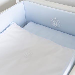 May include: A baby cot with a pale blue bumper and white bedding. The white bedding includes a blanket and a pillow. The bumper and blanket are embroidered with a white monogram design. The cot is white.