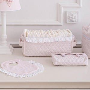 Puede incluir: Un conjunto de accesorios para bebés en rosa pálido. Incluye un cambiador acolchado con ribete de volantes blancos, un babero, un estuche pequeño y un neceser a juego. También hay una lámpara blanca con un lazo rosa.