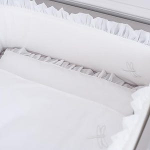Könnte beinhalten: Ein weißes Kinderbett-Set mit einer Rüschenkante am Nestchen und einer passenden Decke. Der Stoff ist sowohl auf dem Nestchen als auch auf der Decke mit dezenten, hellgrau gestickten Libellenmotiven verziert.