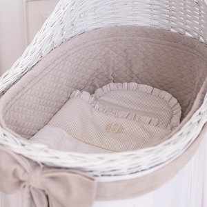 May include: A white wicker bassinet with a soft, quilted interior and a striped pillow. The bassinet features a large bow on the front and is designed for a baby. The bedding is in neutral tones.