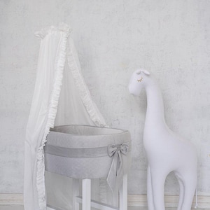 Può includere: Una culla bianca per neonati con interno grigio e fiocco abbinato. Un peluche bianco a forma di giraffa è nelle vicinanze. La culla ha una tenda bianca con bordi a balze. La culla è su un supporto di legno bianco.