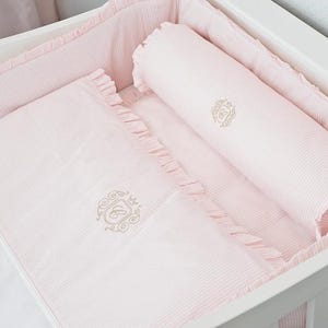 Può includere: Una culla con un set di biancheria da letto a righe rosa e bianche. Il set include un lenzuolo con angoli, un cuscino e un cuscino a rullo, tutti con un emblema ricamato in oro. L'interno della culla è foderato con tessuto abbinato.