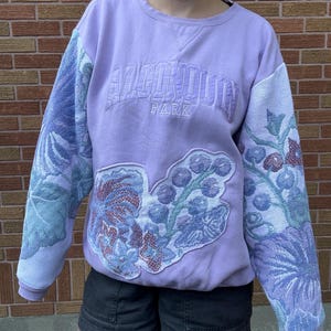 Peut inclure: Un sweat-shirt à col rond violet clair avec le texte brodé "ALGONQUIN PARK" sur la poitrine. Les manches et un grand empiècement frontal présentent un motif floral et de baies tissé style tapisserie dans des tons bleu, vert et rouge.