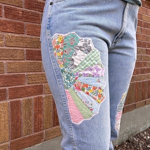 May include: Pale blue denim jeans featuring decorative patchwork fan motifs on the legs. The patchwork incorporates floral, patterned, and solid fabric sections. The jeans have a classic five-pocket design and a straight leg fit.
