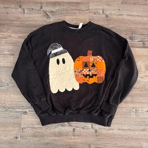 May include: A black crewneck sweatshirt featuring a large applique design on the front. The design includes a cream-coloured ghost wearing a spiderweb-patterned hat next to a patchwork-style orange pumpkin with various Halloween-themed fabric prints.