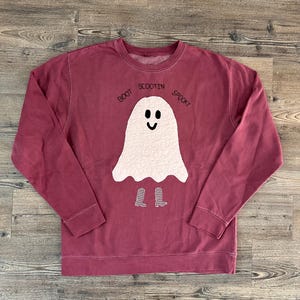 May include: A maroon long-sleeve crewneck sweatshirt featuring a central white ghost appliqué with a quilted texture, black embroidered eyes and mouth, and two small black and white chequered boots. The text "BOOT SCOOTIN SPOOKY" is embroidered in black above the ghost.