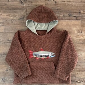 Peut inclure: Un sweat à capuche matelassé marron avec une surface texturée et une doublure de capuche vert clair. L'avant présente une application décorative en forme de poisson faite de divers tissus à motifs, dont des pois rouges et des imprimés floraux.