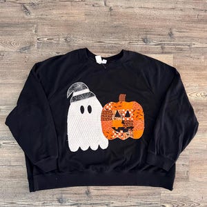 May include: A black long-sleeve sweatshirt featuring a large appliqué design on the front. The design includes a white ghost wearing a spiderweb-patterned hat and an orange pumpkin made of various patterned fabric patches with a black face.