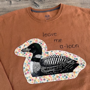 May include: A rust-coloured crewneck sweatshirt featuring an embroidered loon bird appliqué with a floral patterned border. The bird is black and white with a red eye. The text "leave me a-loon" is embroidered in black thread above the bird graphic.