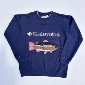 May include: Navy blue sweatshirt with the Columbia Sportswear Company logo and a fish applique. The fish features a patchwork design with red and white gingham fins and a detailed body. The sweatshirt has a crew neck and long sleeves.