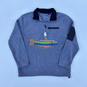 May include: A blue fleece pullover with a black collar and a quarter-zip closure. The front features a colorful fish applique with brown, green, and red accents. A black patch is on the right sleeve. A small tag hangs from the zipper.