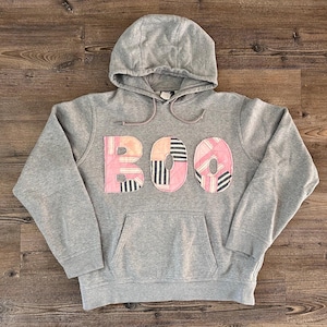 May include: A heather grey hooded sweatshirt featuring the word BOO appliqued on the front. The letters are constructed from quilted fabric patches in shades of pink, white, and black with striped and solid patterns. It includes a front pouch pocket.