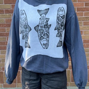 Peut inclure: Un sweat-shirt bleu ardoise à col rond avec un grand empiècement rectangulaire bleu clair sur le devant. L'empiècement présente trois illustrations de poissons à l'encre noire, ornées de motifs floraux complexes sur tout le corps.