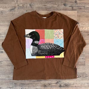 May include: A brown long-sleeve shirt featuring a central rectangular patch with a patchwork grid design. The patch displays a black and white illustration of a loon bird set against a background of various colourful patterned fabric squares.