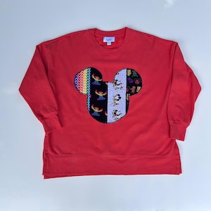 May include: A red sweatshirt featuring a Mickey Mouse head design. The Mickey Mouse head is made of colourful patches with various patterns, including rainbows, Stitch, and Mickey Mouse. The sweatshirt has a crew neck and long sleeves.