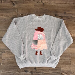 May include: A heather grey crewneck sweatshirt featuring a central appliqué of a ghost character. The ghost is constructed from pink and black-and-white striped fabric patches, wearing a brown cowboy hat and matching brown cowboy boots.