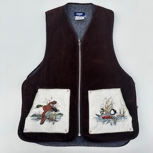 May include: A brown corduroy vest with a full-length zip and two front pockets. The pockets have embroidered images of ducks in a wetland scene. The vest has a grey fleece lining and a small tag near the neckline.