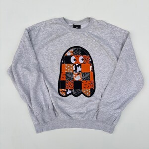 May include: A pale grey sweatshirt featuring a Halloween ghost design. The ghost is a patchwork of orange, black, and white patterned fabrics. The ghost has two large, round eyes.