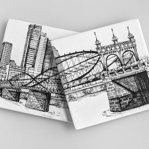 May include: Two black and white ink drawings on ceramic coasters. One coaster features a cityscape with a bridge and skyscrapers. The other coaster features a bridge with a detailed design.