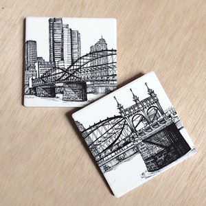 May include: Two black and white ceramic coasters featuring ink drawings of bridges in a city skyline. The first coaster shows a bridge with a cityscape in the background. The second coaster shows a close-up of a bridge.