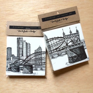 May include: Set of two white ceramic coasters with black ink drawings of bridges in Pittsburgh, Pennsylvania. The coasters are packaged together with a brown paper label that reads "Pittsburgh, art coaster set "Smithfield st Bridge""