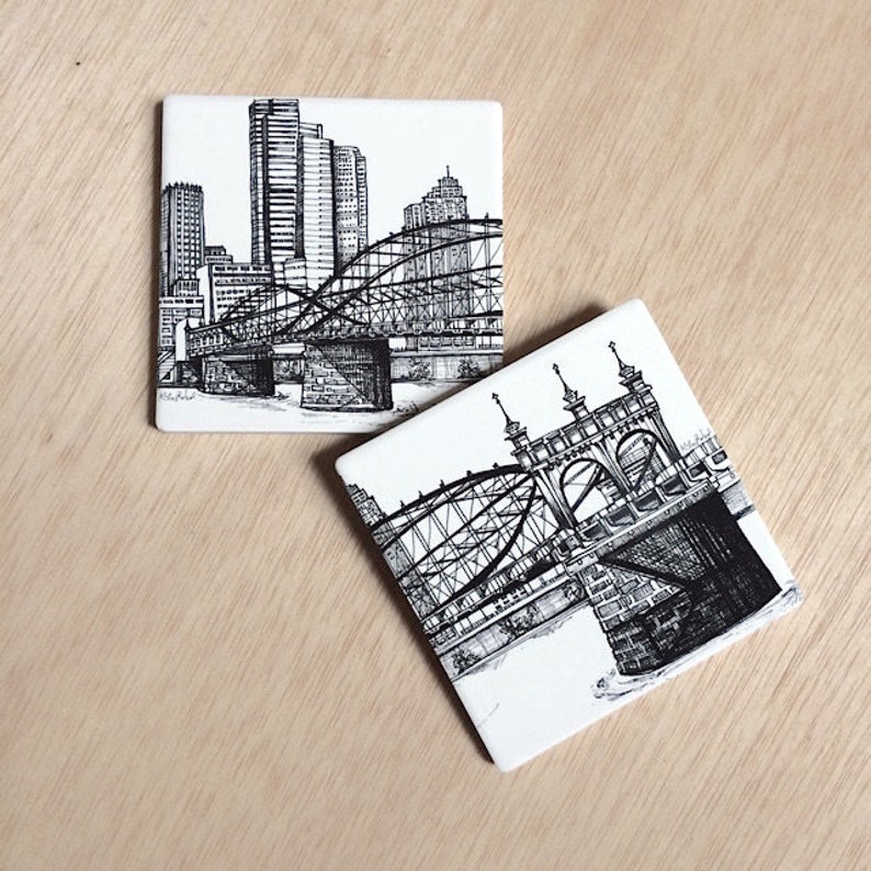 May include: Two black and white ceramic coasters featuring ink drawings of bridges in a city skyline. The first coaster shows a bridge with a cityscape in the background. The second coaster shows a close-up of a bridge.