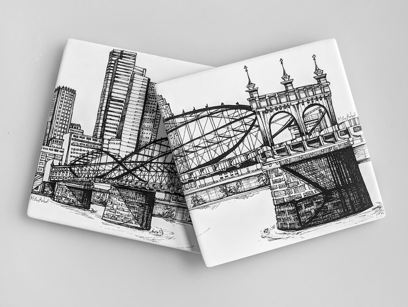 May include: Two black and white ink drawings on ceramic coasters. One coaster features a cityscape with a bridge and skyscrapers. The other coaster features a bridge with a detailed design.