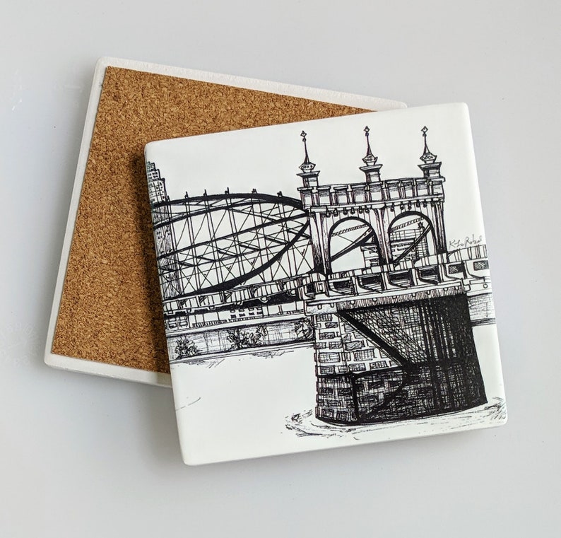 May include: Black and white line drawing of a bridge with a detailed design. The bridge is made of metal and has a series of arches. The drawing is on a white ceramic coaster with a cork backing.